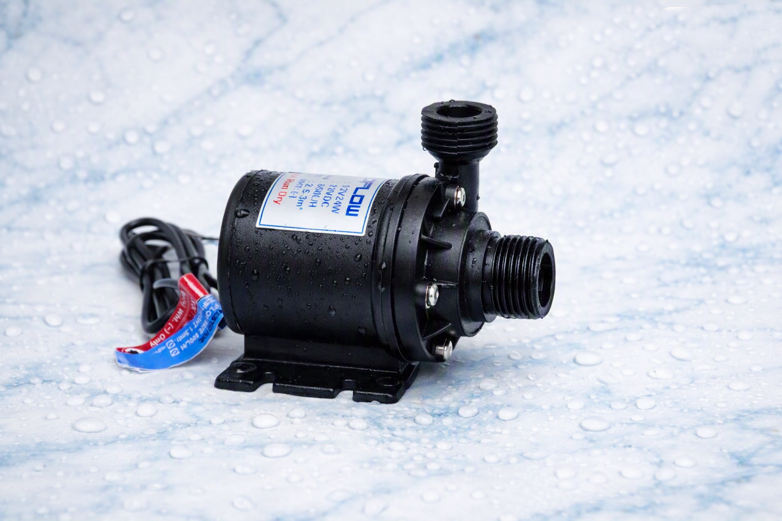 Compact black 12V DC water pump with cylindrical body, two threaded pipe connectors, and red/blue wires, placed on a wet surface with visible water droplets.