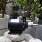 Zeddflow 12V DC submersible water pump resting on white stones with stainless steel G1/2 inch female barb adapters installed.